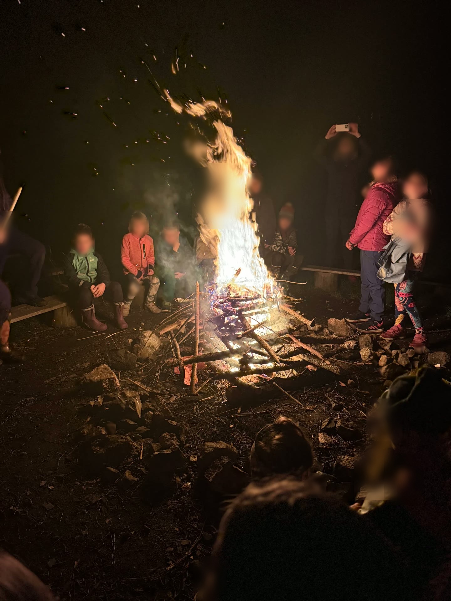 Děti a dospělí stojí a sedí v kruhu u velkého ohně během večerního rituálu